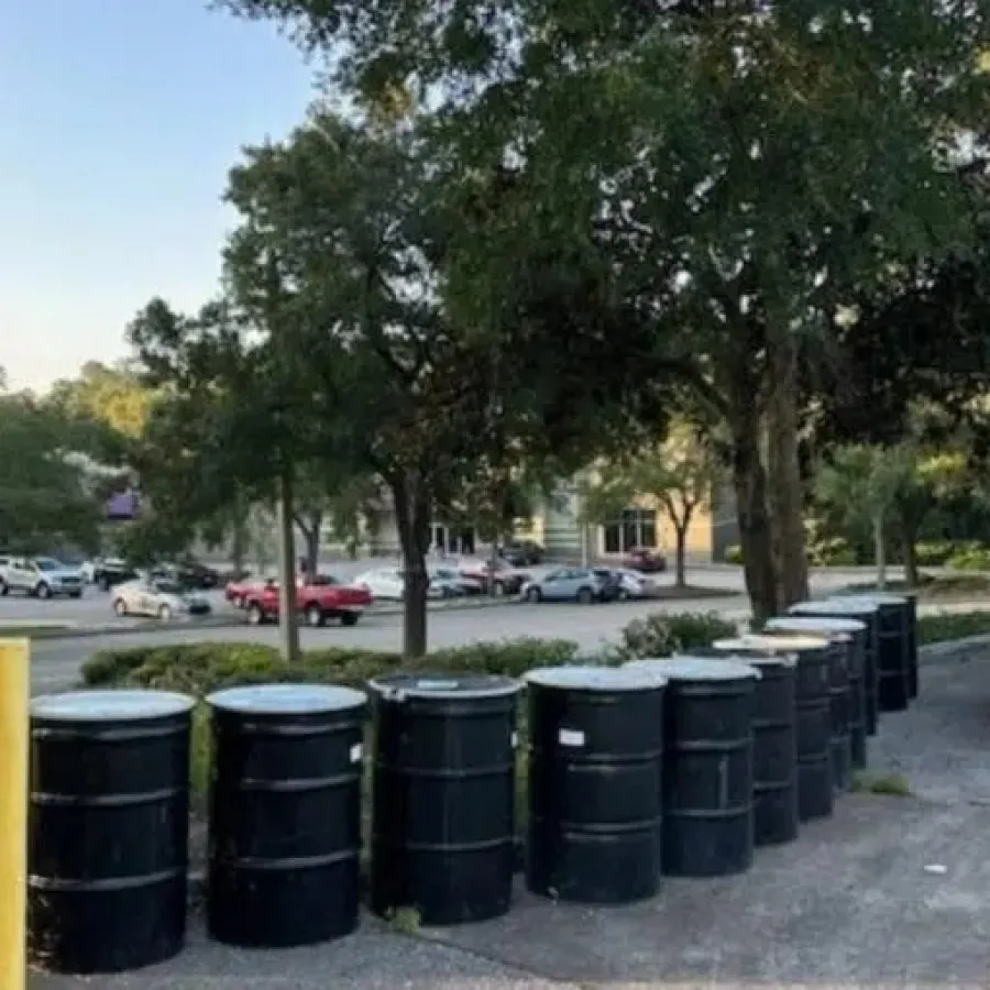 Row of black metal barrels lined up along a parking lot edge with trees and cars in the background