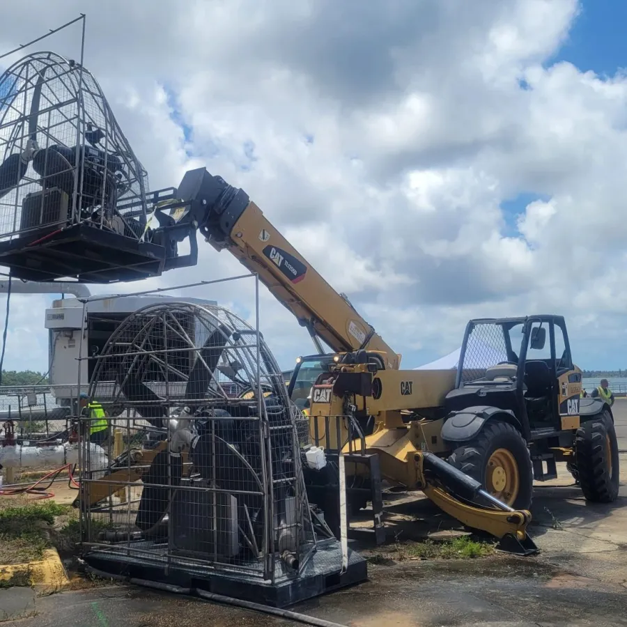 Construction equipment lifting large industrial fan cages with workers and vehicles in an outdoor industrial area under blue sky.