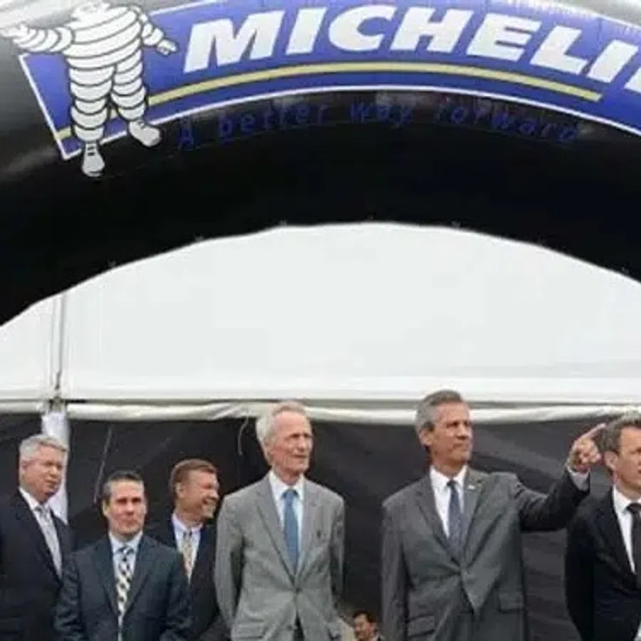 Group of six men in suits standing under a large black Michelin branded arch at a formal event.