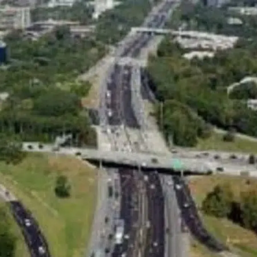 Aerial view of a busy highway intersection surrounded by greenery and urban buildings in the background.