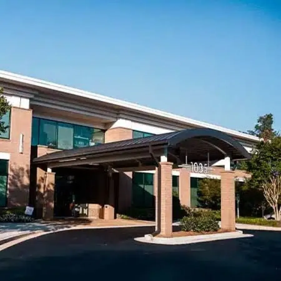 Modern two-story office building with large windows, shaded entrance, and landscaped trees and bushes.