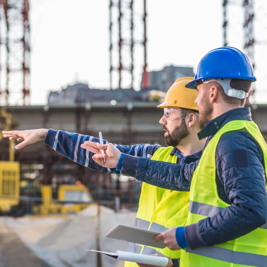 Two construction workers wearing helmets and safety vests discuss plans at a building site with a tablet and blueprints.