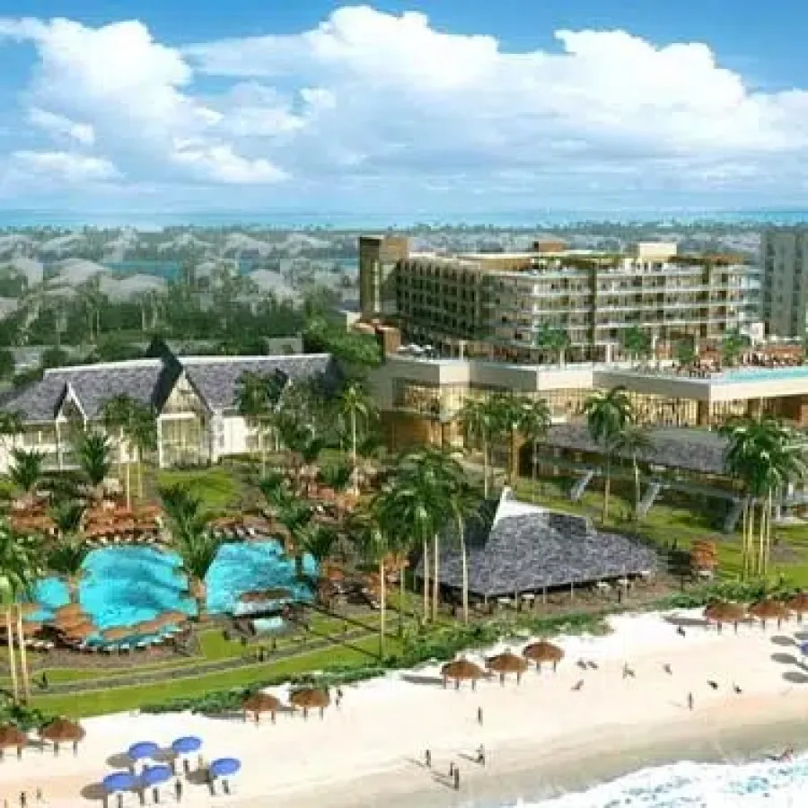 Aerial view of a beachfront resort with pools, palm trees, umbrellas, and modern buildings near the ocean shore under blue skies.