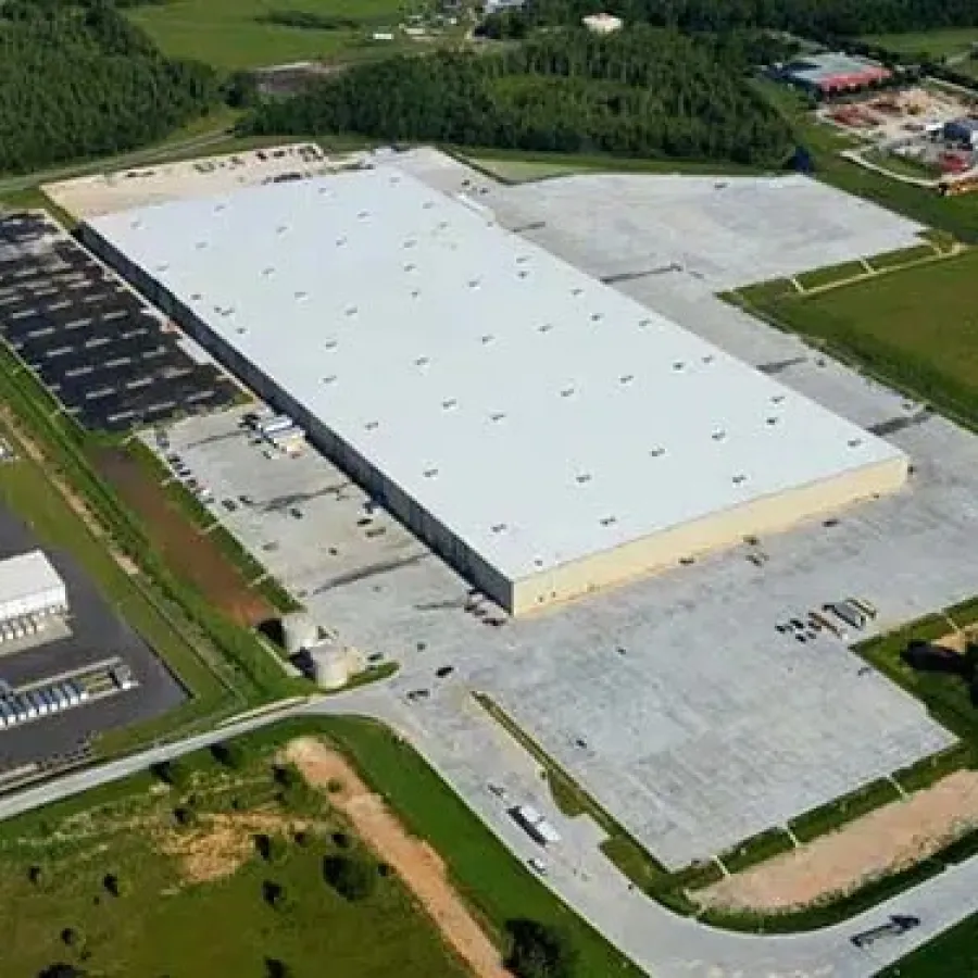 Aerial view of a large industrial warehouse with adjacent parking lots surrounded by green fields and roads.