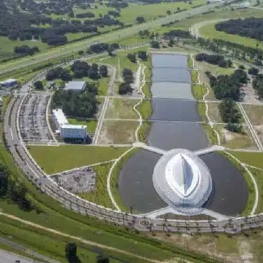Aerial view of a futuristic circular building surrounded by water and greenery with adjacent parking areas and highways.