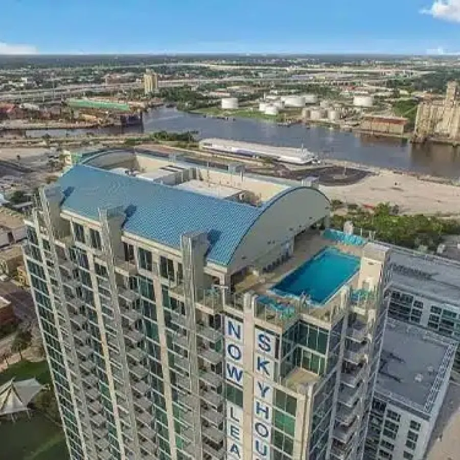 Aerial view of NOWLAR Skyhouse high-rise building with rooftop pool near river and urban landscape.