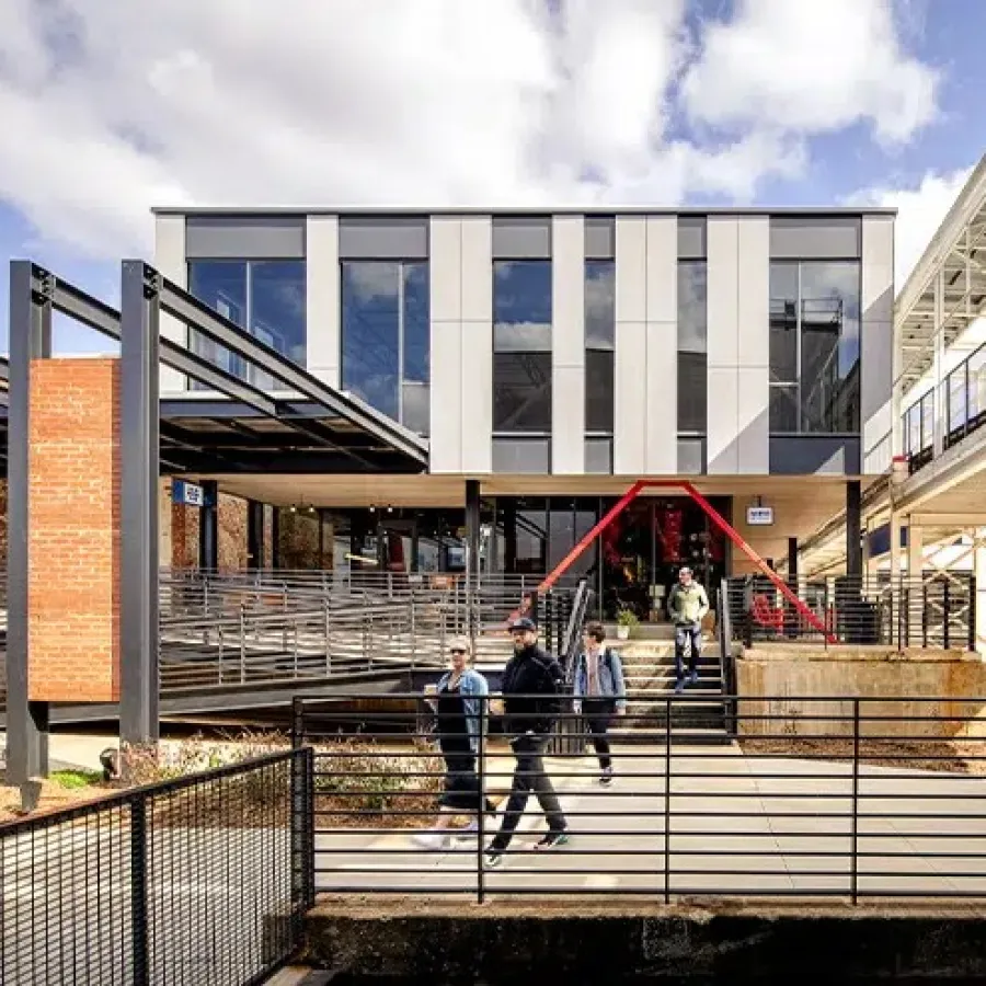Modern industrial building with metal beams, brick walls, and people walking near railing on sunny day.