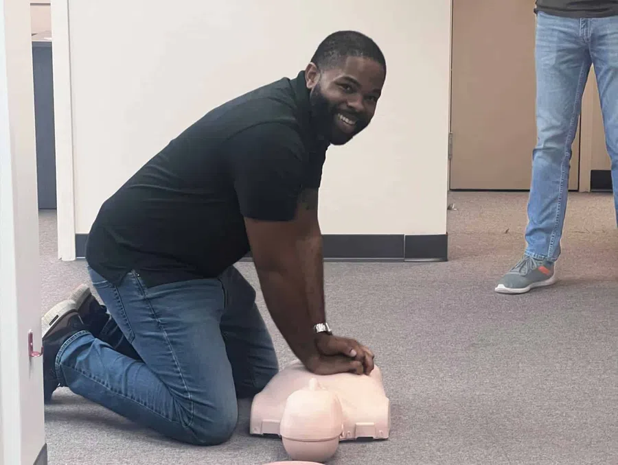 Man kneeling on the floor performing CPR on a training mannequin in an indoor setting.