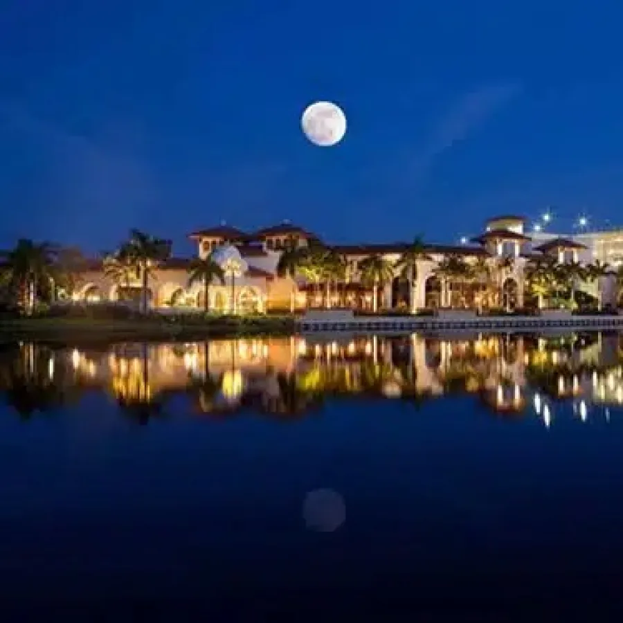 Luxurious resort illuminated at night with full moon reflecting on calm lake water under clear sky.