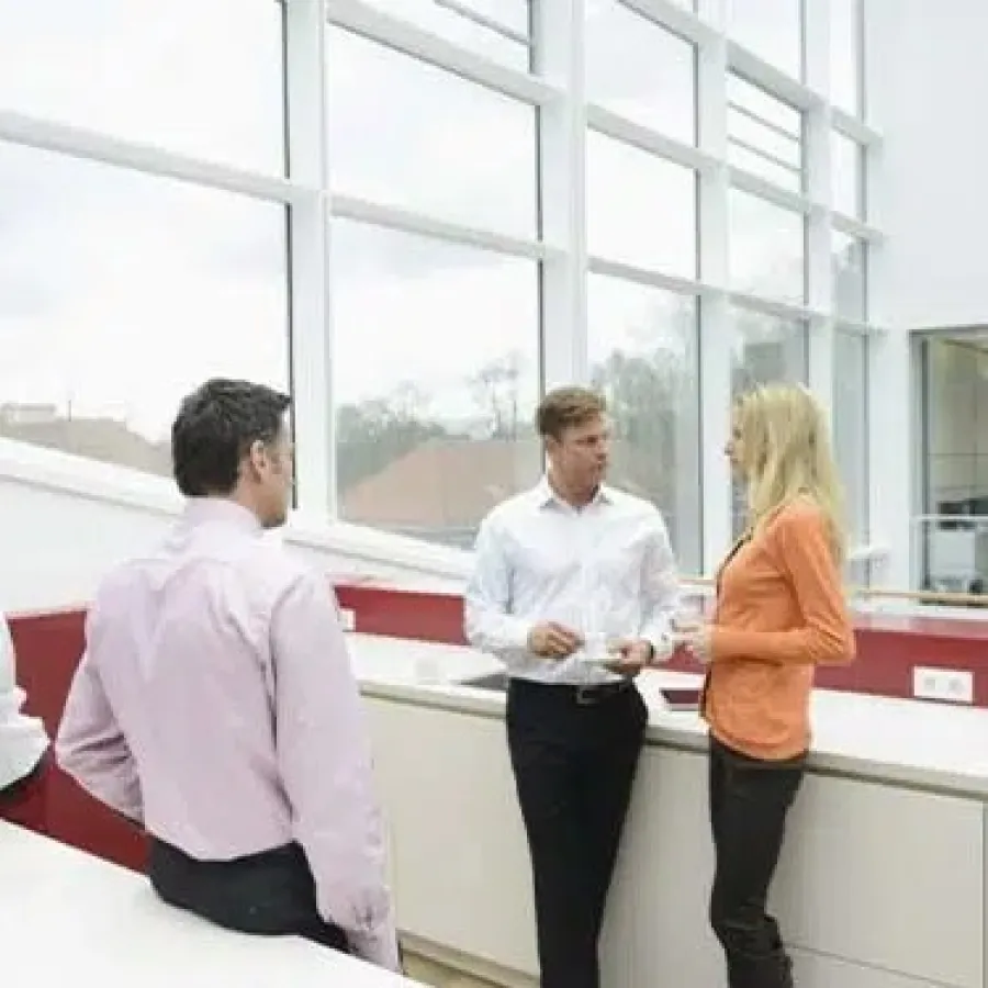 Four professionals interacting in a modern office break area with large windows and natural light.