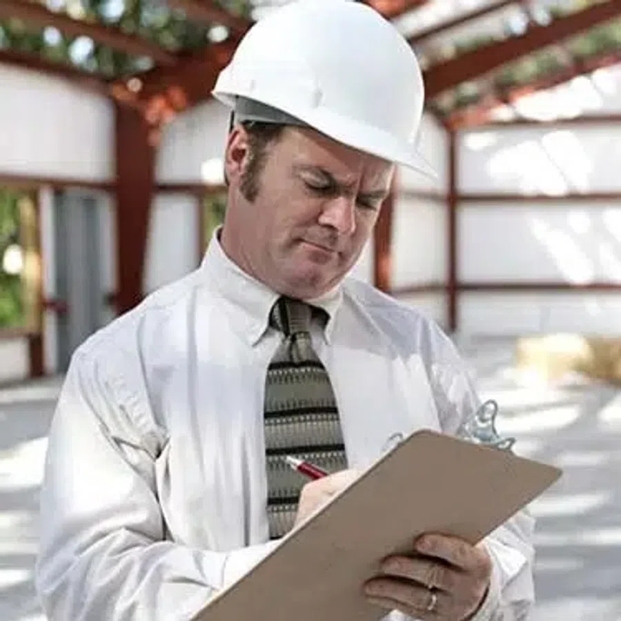 Engineer in white hard hat and tie inspecting and writing notes on clipboard at construction site.