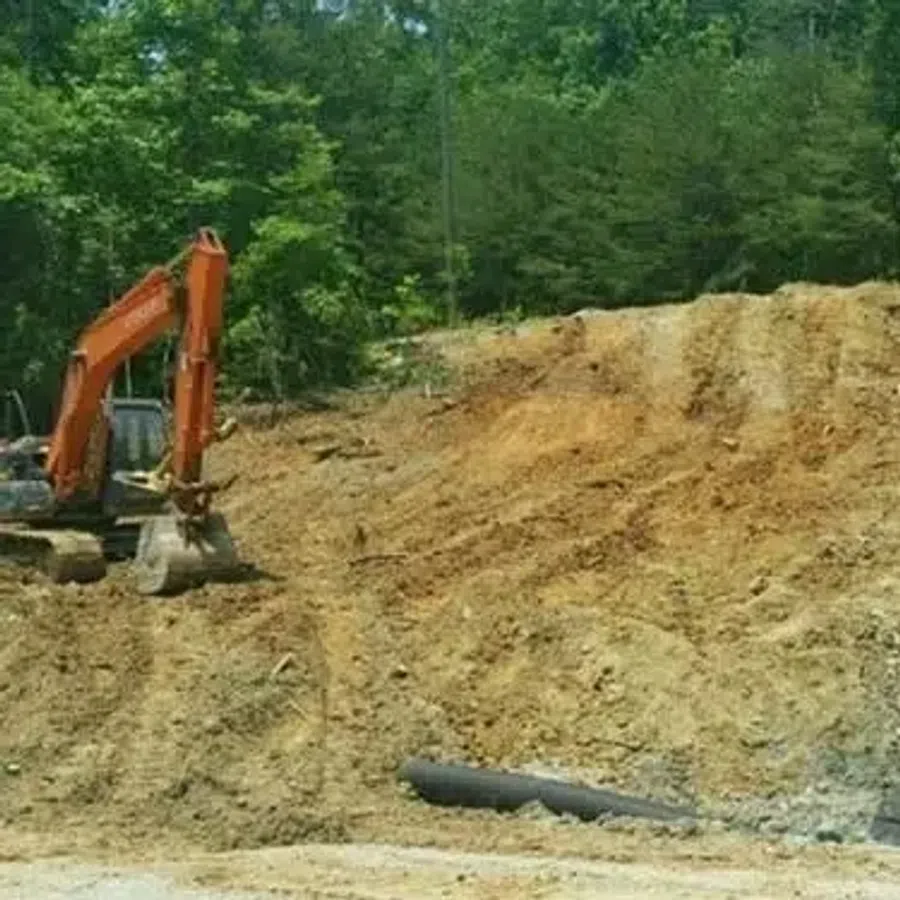 Excavator beside a large dirt slope with black erosion control fabric installed in a wooded area.