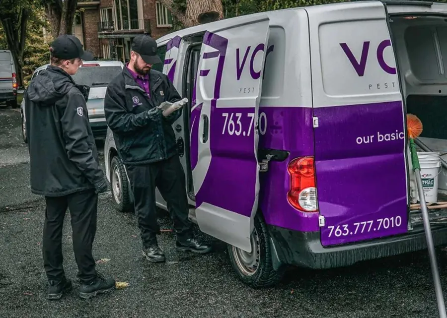 Two pest control workers in uniform consulting near a white and purple service van with equipment inside.