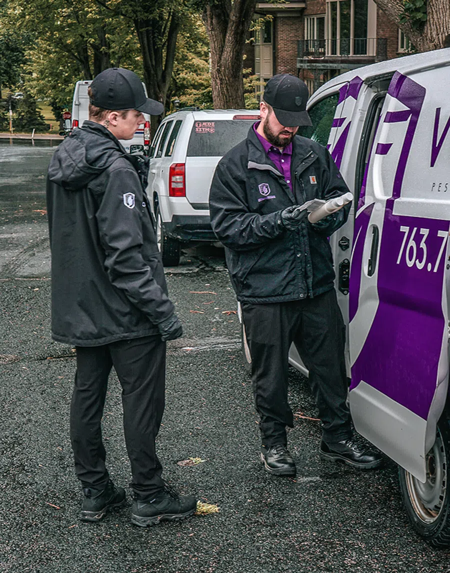 Two pest control workers in black uniforms checking equipment beside a branded white and purple service van.