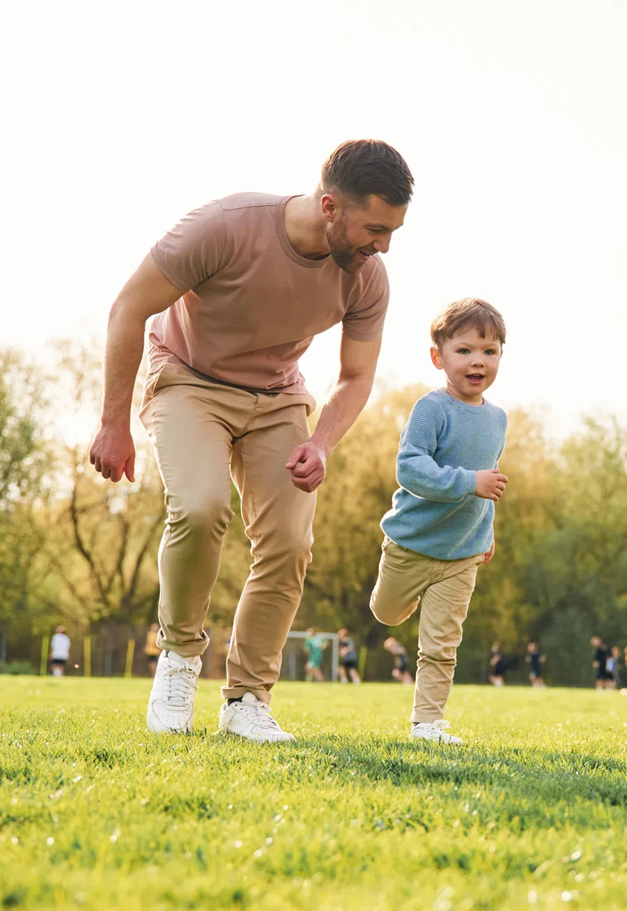 Father and young son running and playing happily on a sunny grass field in the park with trees in background