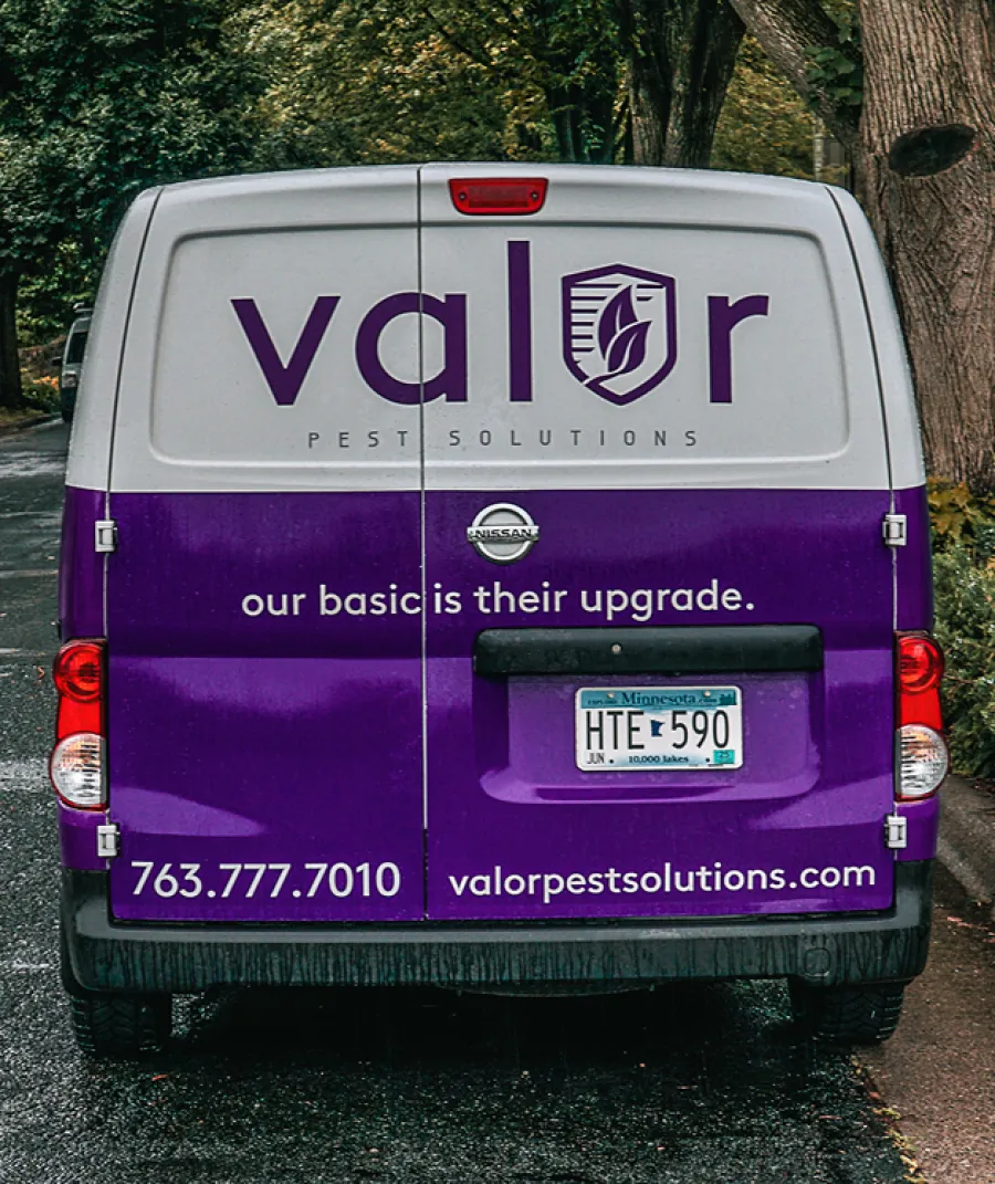 Rear view of a purple and white Nissan van with Valor Pest Solutions branding and contact details on a tree-lined road.