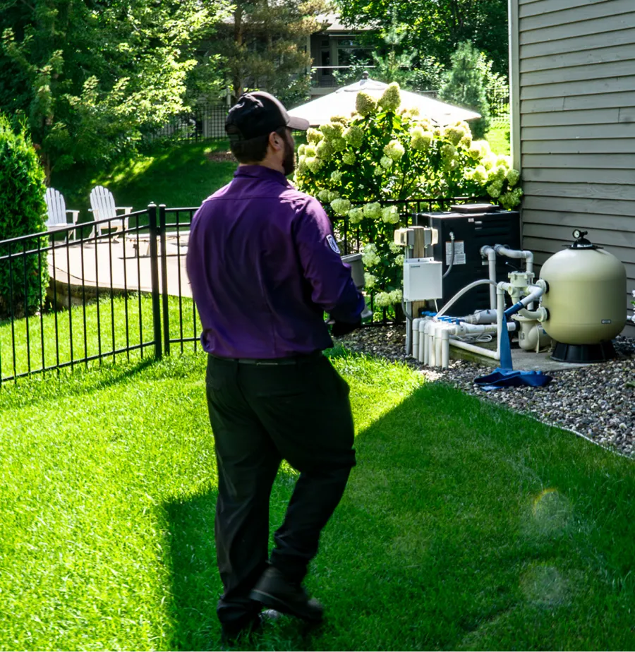 Technician in purple shirt and cap inspecting backyard pool equipment near a house in bright sunlight.