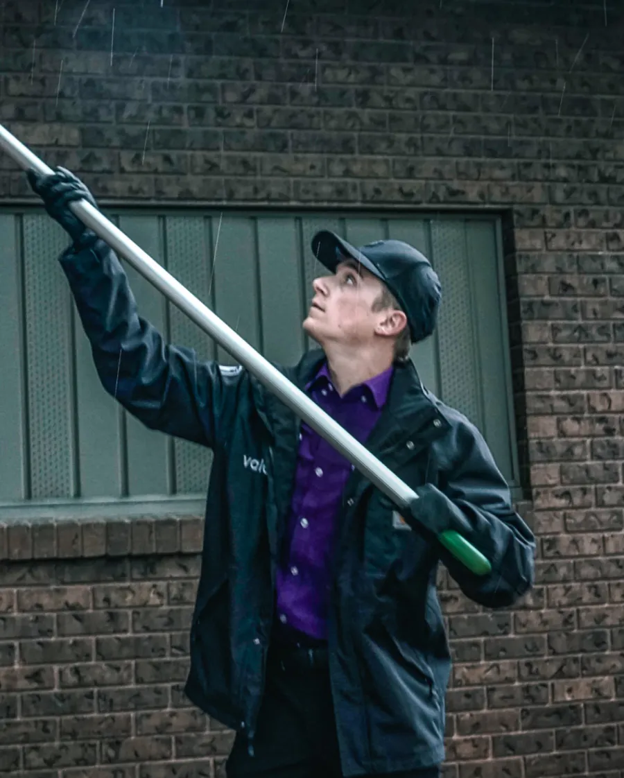 Man in black jacket and cap holding a long pole, looking upward against a rainy brick wall background.