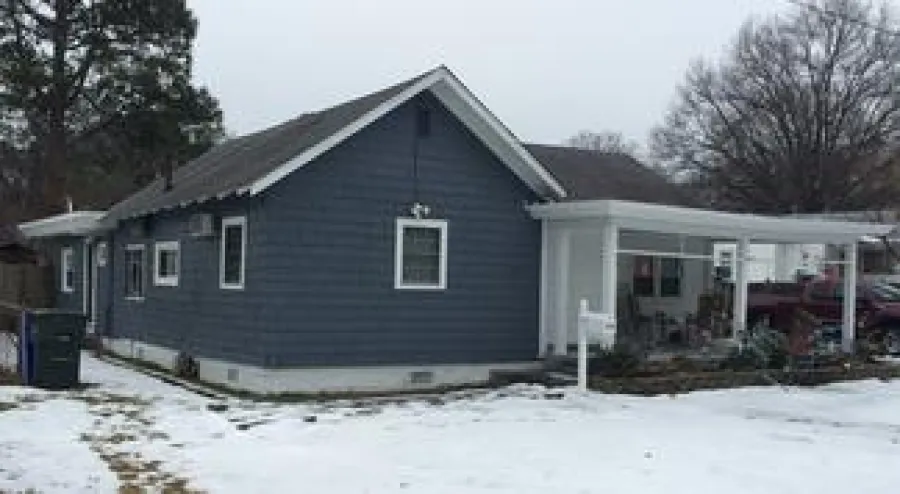 Blue single-story house with white trim surrounded by snow and leafless trees on a cloudy day