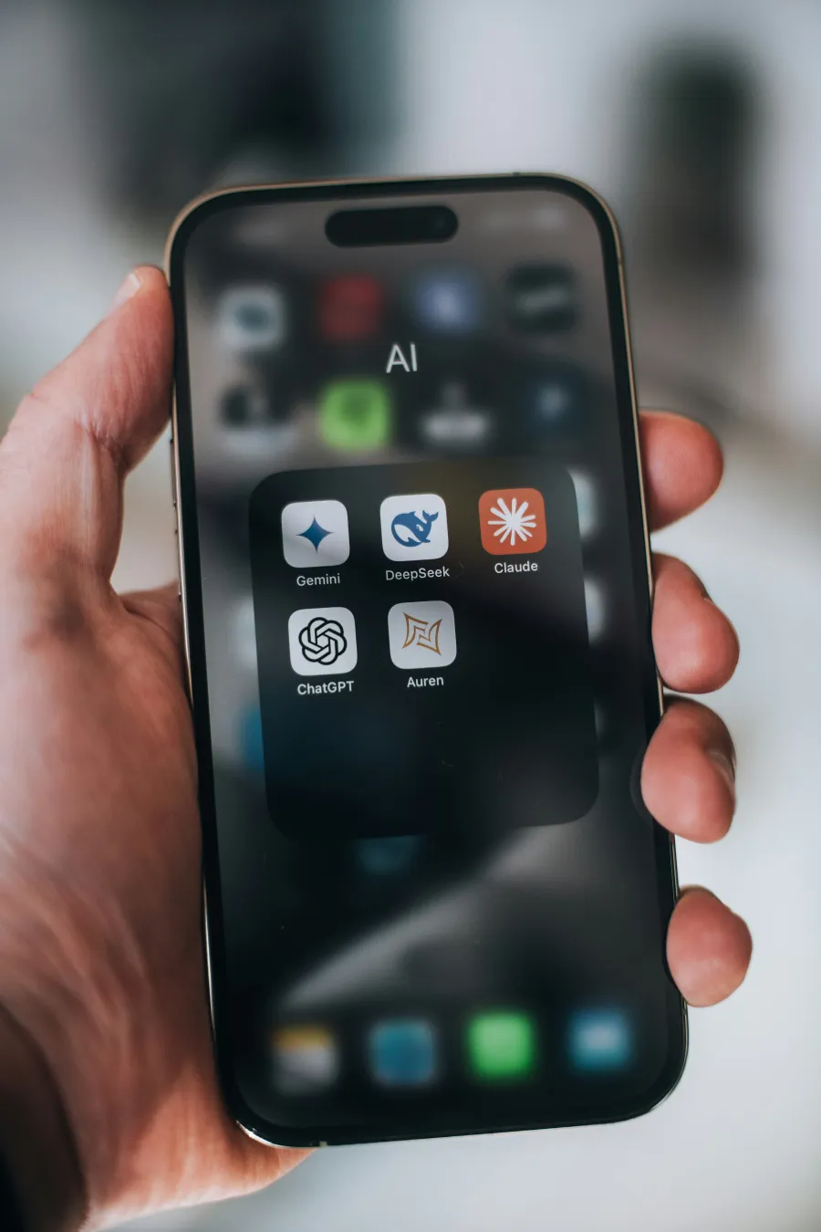 Hand holding smartphone showing AI app folder with Gemini, DeepSeek, Claude, ChatGPT, and Auren icons.