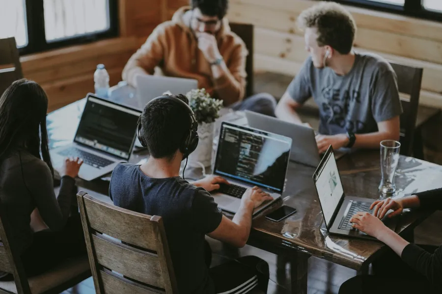 Group of young professionals working on laptops in a casual modern office setting with wooden walls.