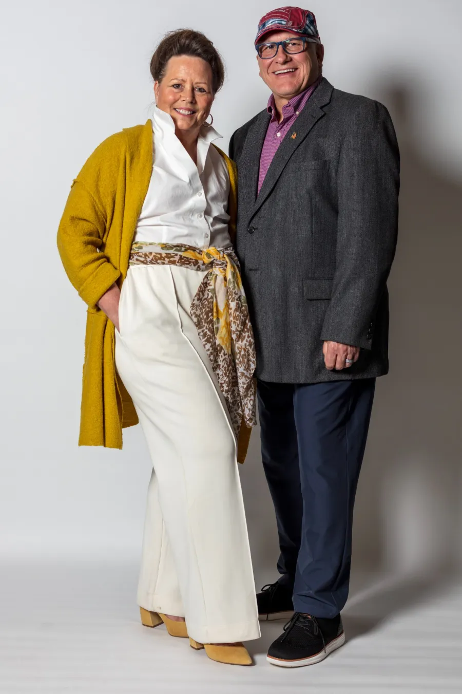 Smiling older couple dressed in stylish formal and casual outfits standing against a white background.