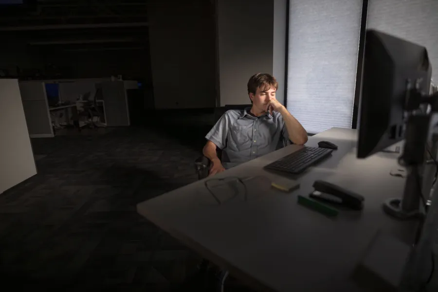 Man in gray shirt sitting alone at desk with computer in a dimly lit office space looking contemplative.