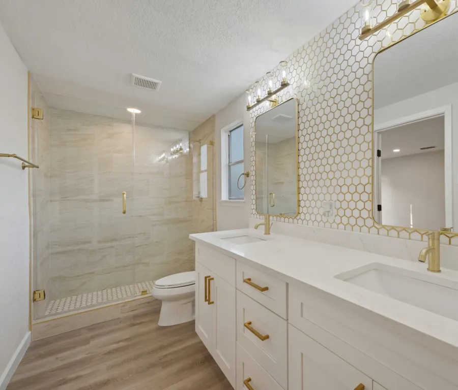Modern white bathroom with double vanity, gold fixtures, hexagonal wall tiles, and glass-enclosed shower.