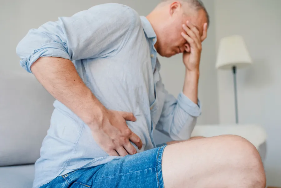 A man in a light shirt clutches his stomach, visibly in pain, seated on a couch at home.
