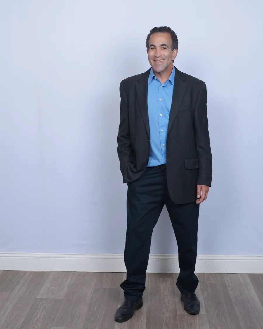Man in dark blazer, blue shirt, and black pants standing on wood floor against a plain light gray background.