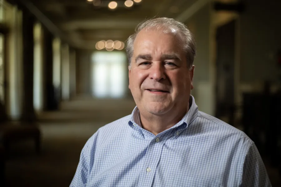 Middle-aged man in a checkered shirt smiling softly in a softly lit indoor hallway with blurred background.