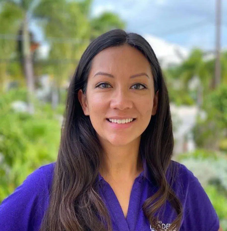 Smiling woman with long dark hair wearing a purple polo shirt outdoors with greenery and sky in background.