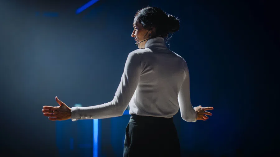 Woman speaking on stage with headset microphone, gesturing with hands under spotlight on dark background