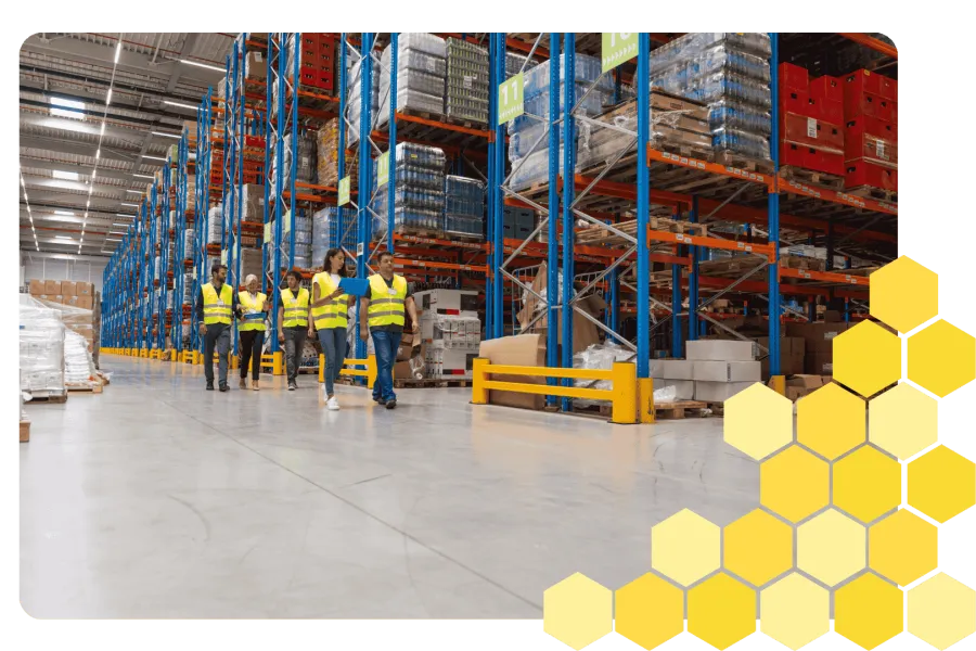 Warehouse workers in yellow vests walking through aisles with tall shelves stocked with boxes and pallets.