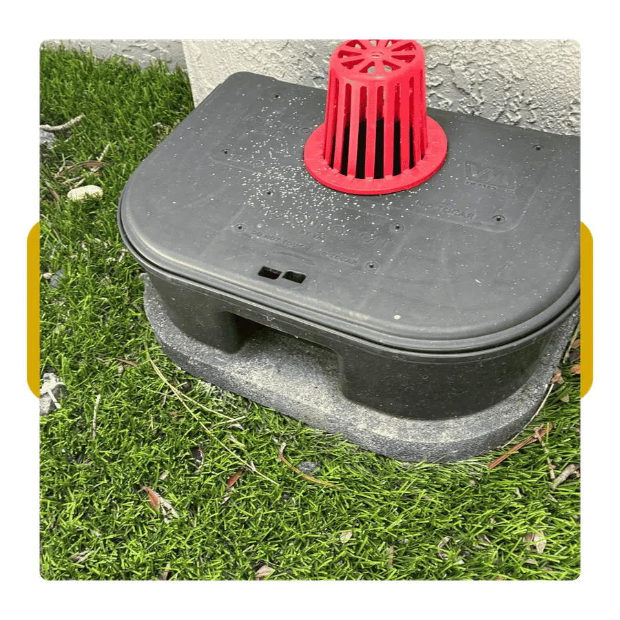 Black irrigation valve box with a red vent cap installed on green grass next to a white wall corner.
