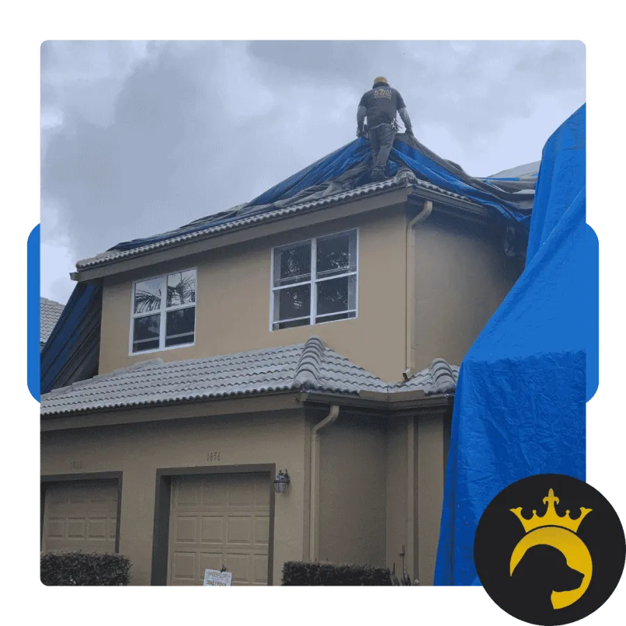 Worker on roof covering tiles with blue tarp on a two-story house during cloudy weather for repairs.
