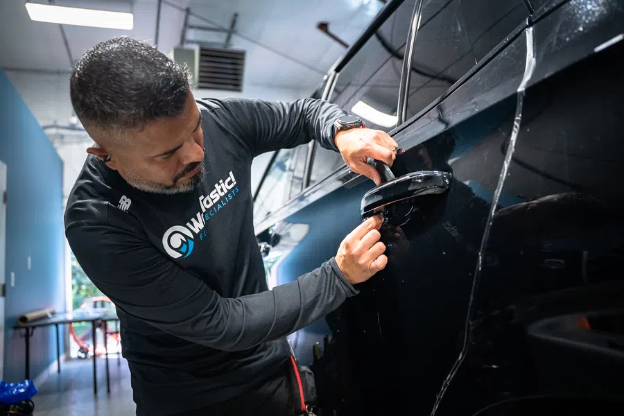 Technician with WFASTICS shirt repairing or installing a door handle on a black car in a service garage.