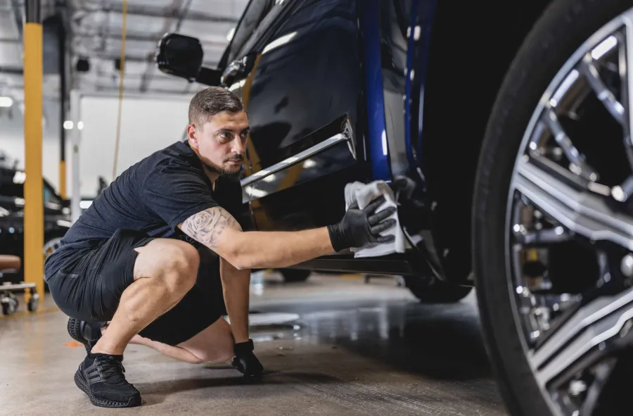 Man wearing black gloves carefully polishing a dark blue car in a well-lit garage.