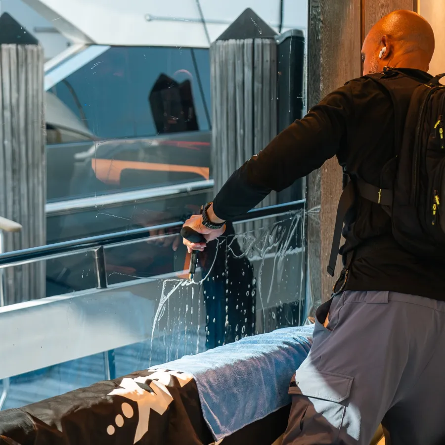 Man cleaning yacht window with squeegee on dock, wearing black jacket and backpack, reflected in glass
