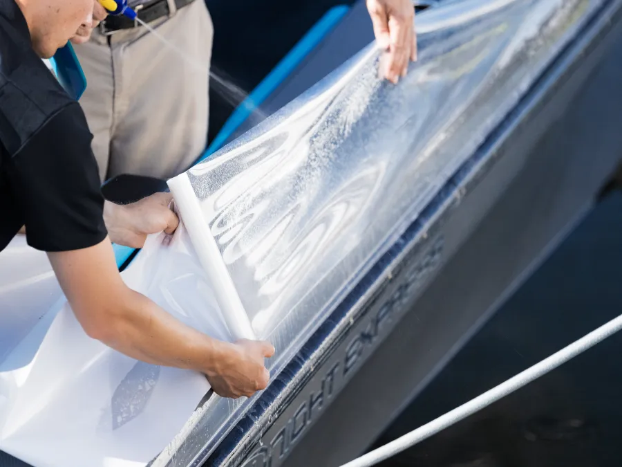 Two people carefully applying protective film onto a boat surface near the water.