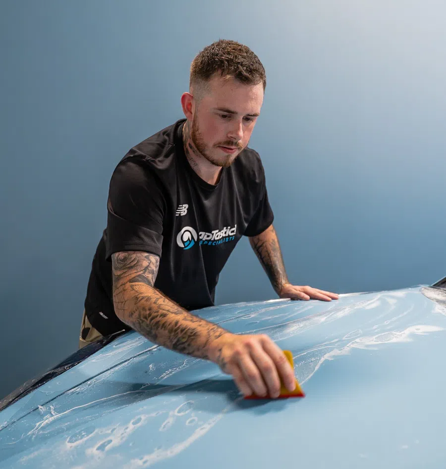Man with tattoos carefully applying protective film on a car hood using a squeegee in a workshop.