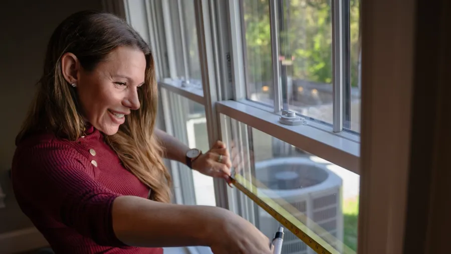 Woman in red sweater measuring window width with a tape measure indoors on a sunny day.