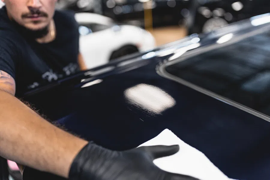 Person wearing black gloves polishing a shiny black car surface with a white cloth inside a garage.