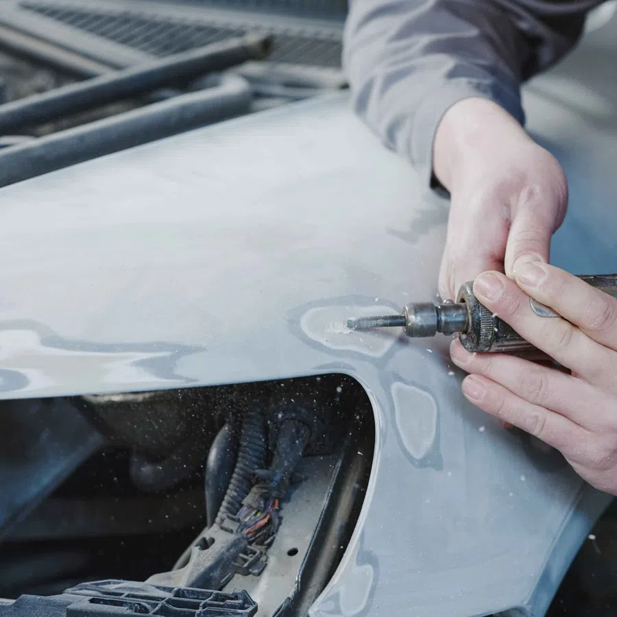 Hands using a rotary tool to repair and smooth a car's body panel near an engine compartment opening.